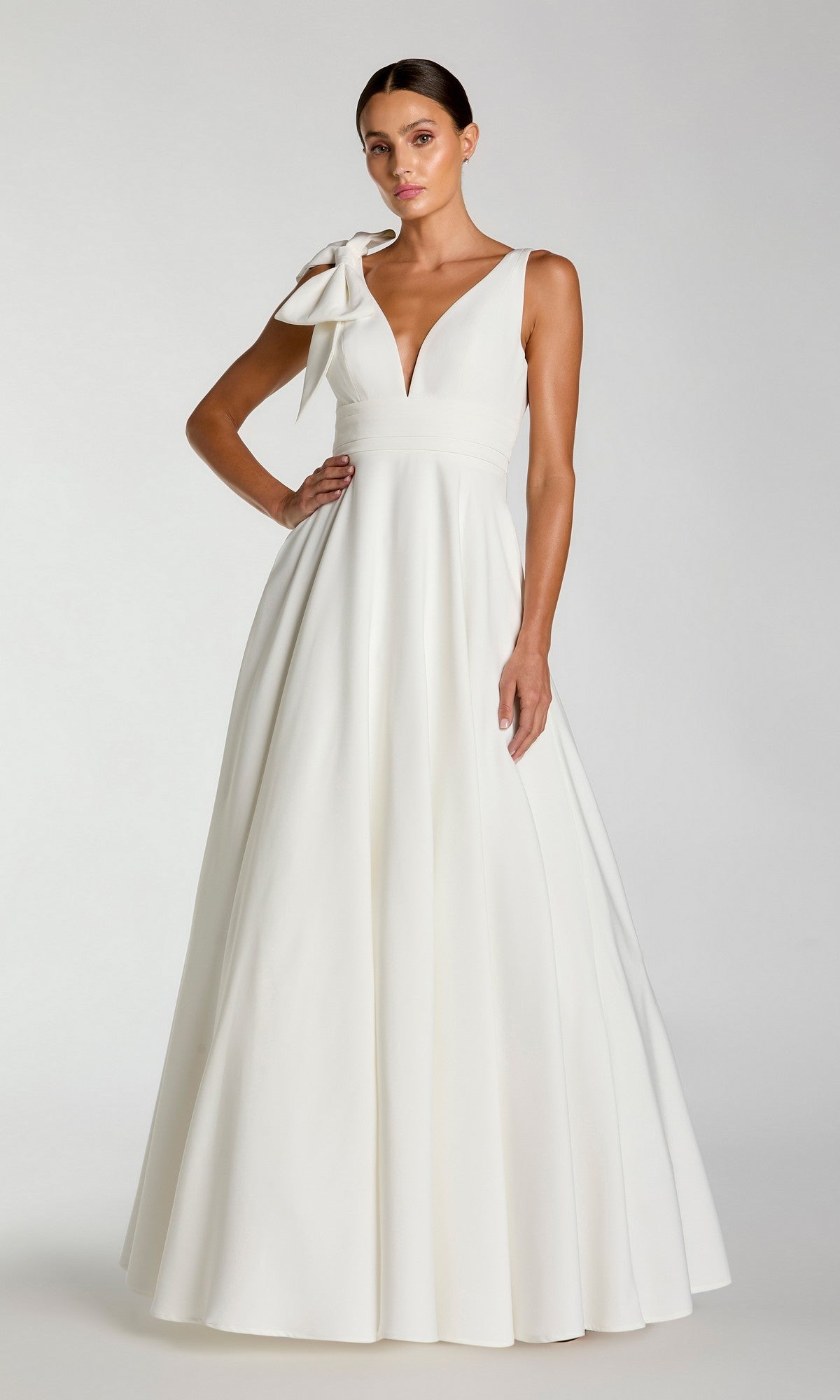A woman stands against a plain light background wearing the Mac Duggal 49972 Shoulder-Bow Long Formal Dress, featuring an elegant sleeveless design, deep V-neckline, and a large bow accent on one shoulder.